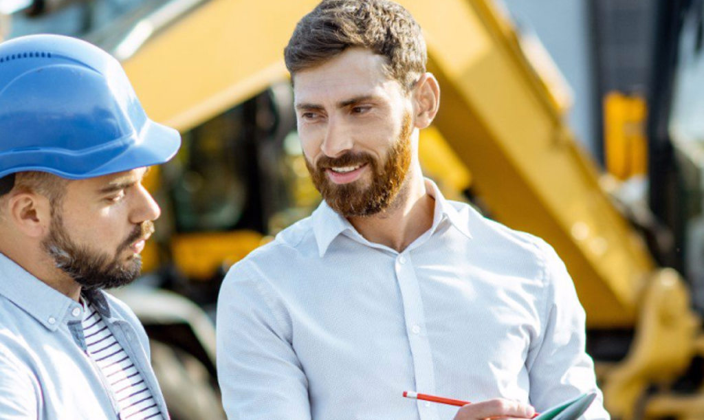 This image depicts a business owner talking to an employee who is wearing a blue hard hat while they stand in front of yellow construction equipmment.